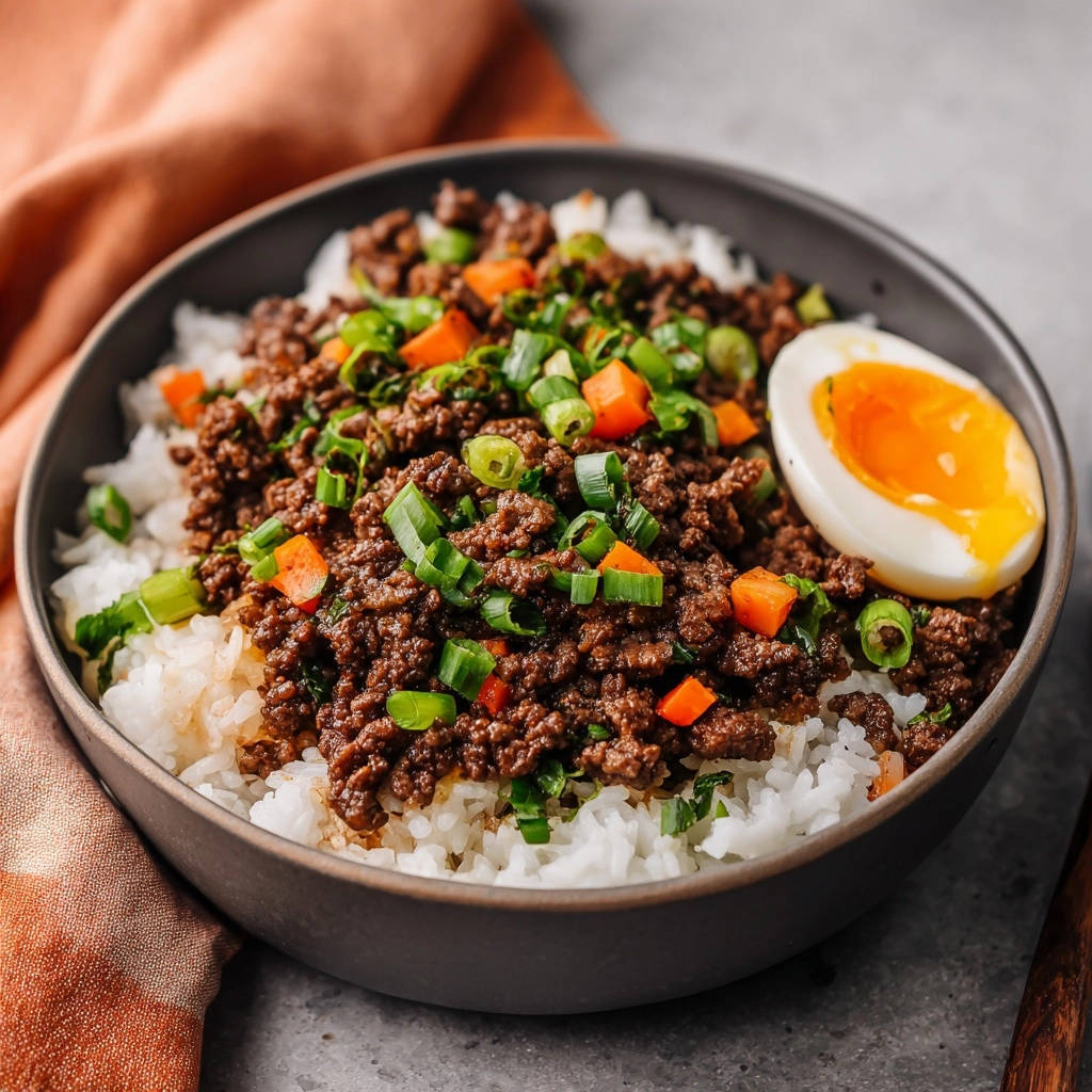 Würzige Hackfleisch-Bowl mit Spiegelei serviert in einer dunklen Schüssel, garniert mit frischen Kräutern und Karottenwürfeln.