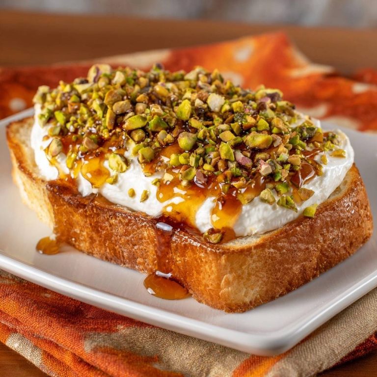 Knuspriger Pistazien-Ricotta-Toast, serviert auf einem weißen Teller, mit geschmolzenem Honig, der an den Seiten herunterläuft und mit gehackten Pistazien bedeckt ist.